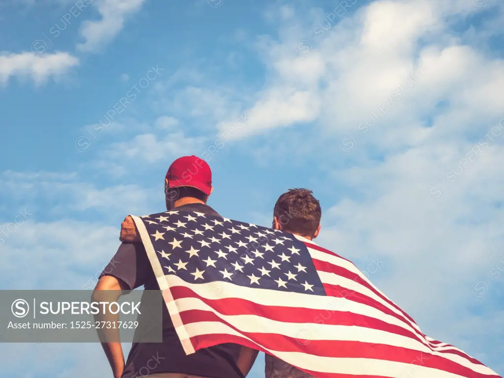 Two men holding Flag of the United States on blue sky background on a clear, sunny day. View from the back, close-up. National holiday concept. Two men holding Flag of the United States