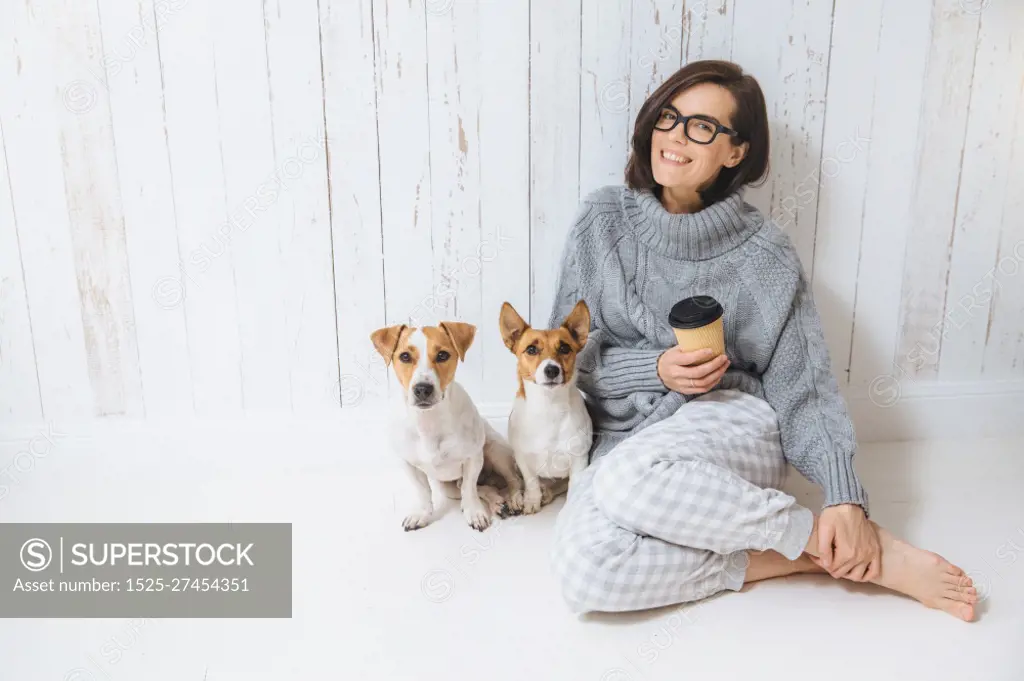 Smiling cheerful young female wears warm woolen sweater, square eyewear, drinks hot beverage, relaxes on floor and her two favourite pets, look directly into camera. People and animals conecpt