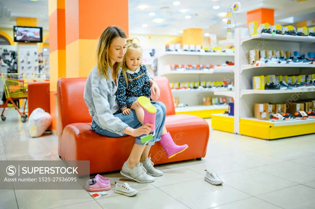 Mother and little daughter buying shoes in kid&rsquo;s store. Mom and adorable girl near the showcase in children&rsquo;s shop, happy childhood, family makes a purchase in market. Mother and little daughter buys shoes, kid&rsquo;s store