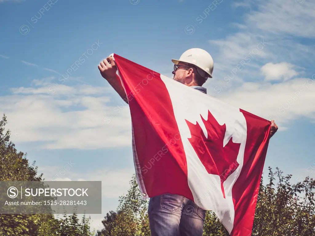 Handsome engineer, holding Canadian Flag in the park against the backdrop of green trees and the setting sun, looking into the distance. Close-up. Concept of labor and employment. Engineer, holding Canadian Flag in the park