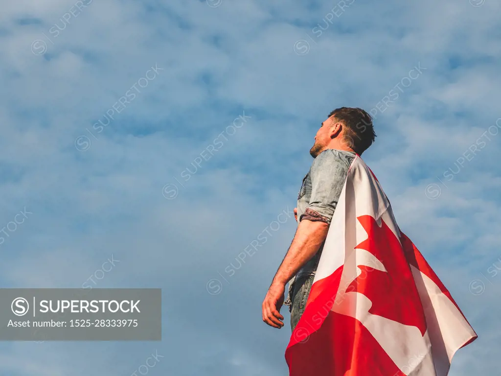 Attractive man in jeans and denim shirt holding a Canadian Flag against a clear, sunny, blue sky. View from the back, close-up. National holiday concept. Man holding a Canadian Flag. National holiday