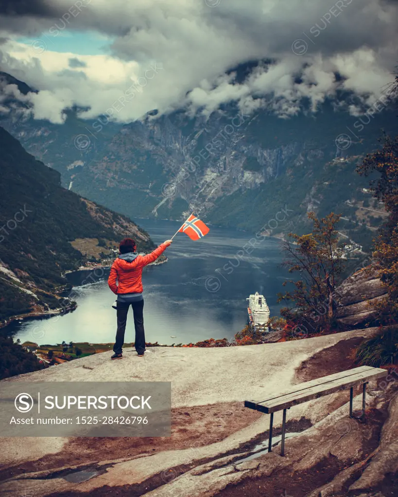 Female tourist enjoying scenic view over fjord Geirangerfjorden from Flydalsjuvet viewpoint, holding norwegian flag. Cruising vacation and travel.. Tourist over Geirangerfjord holds norwegian flag