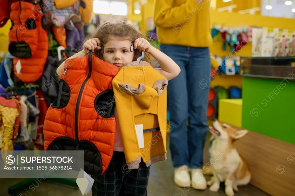 Little girl posing with trendy clothes for dog over pet shop interior. Happy mother and daughter shopping together buying new apparel for their corgi dog. Little girl posing with trendy clothes for dog over pet shop interior