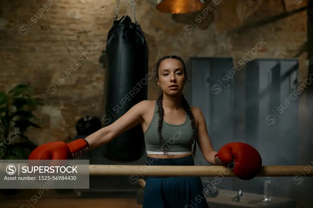 Athletic female fighter with red boxing gloves standing leaned at wooden training bar in gym with loft interior design. Athletic female fighter with bandaged hands standing leaned at wooden bar