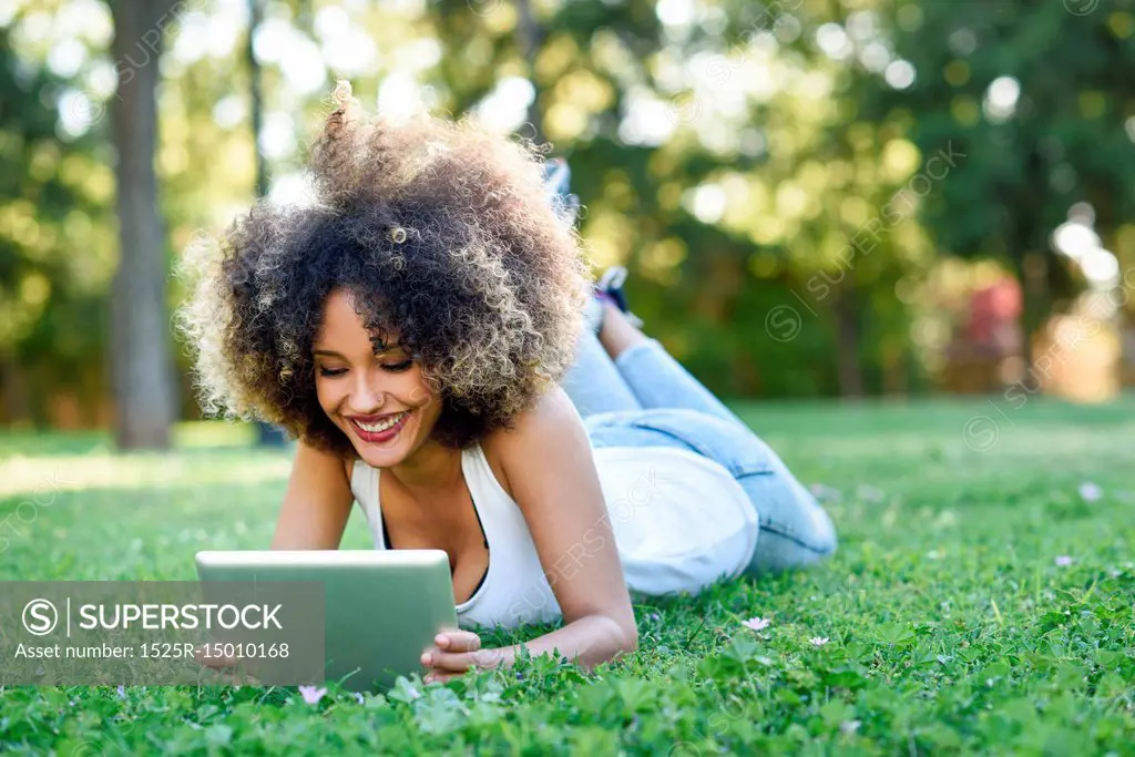 Young black woman with afro hairstyle laying on grass in urban park looking at her tablet computer. Mixed girl wearing casual clothes.