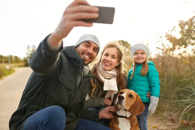 family, pets and people concept - happy mother, father and little daughter with beagle dog taking selfie by smartphone outdoors in autumn. happy family with dog taking selfie in autumn