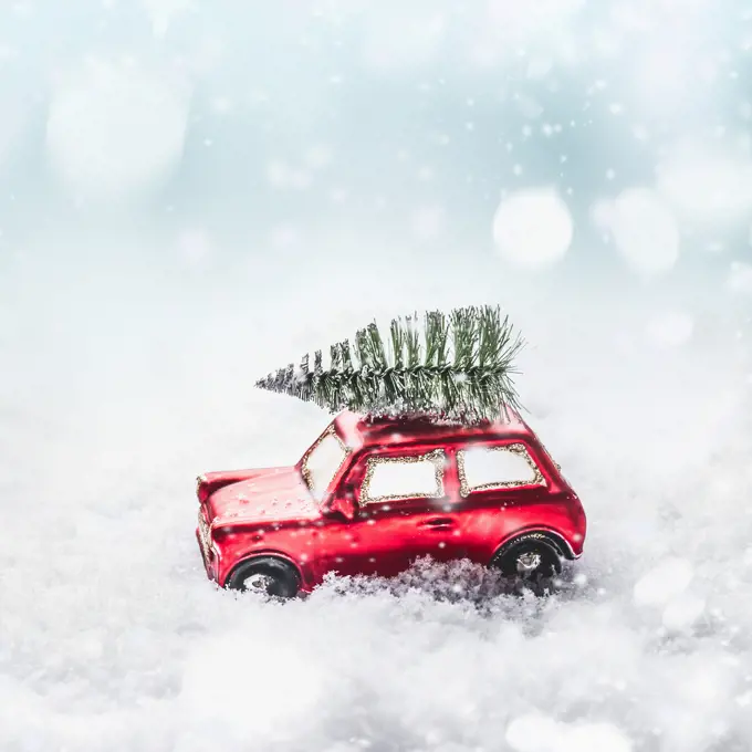 Christmas tree on red toy retro car in snow, through snowy winter wonder land with snowfall and bokeh. Creative Christmas holiday concept. Copy space for your greeting and design. 