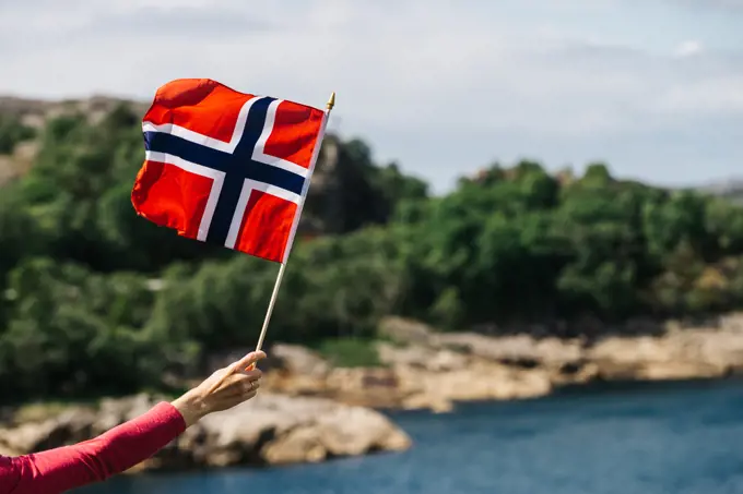 Tourist hand holding norwegian flag on rocky stone sea coast background.. Tourist with norwegian flag on sea coast