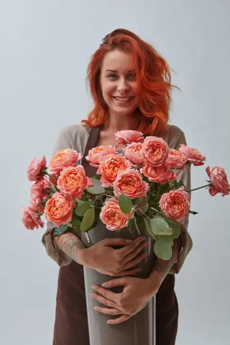 A smiling red-haired woman in a brown apron holds a vase with roses in a color of the year 2019 Living Coral Pantone on a gray background with space for text. The concept of a flower shop. Young woman with a big vase of roses flowers in a color of the year 2019 Living Coral Pantone on a gray background with copy space.