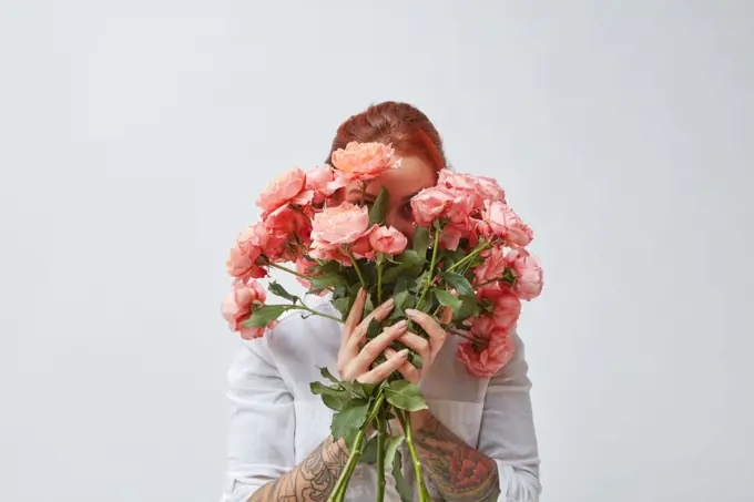 A happy woman with a tattoo on her hands hides her face behind a bouquet of fresh roses in a color of the year 2019 Living Coral Pantone. Postcards for Mother&rsquo;s Day.. Congratulation bouquet of roses in the gerl&rsquo;s hands with a tattoo on a white background. Trend color of the year 2019 Living Coral Pantone.