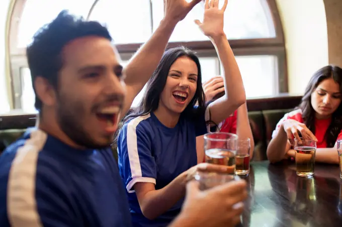 sport and entertainment concept - happy football fans or friends drinking beer, making high five and celebrating victory at bar or pub, supporting two teams with different shirt color