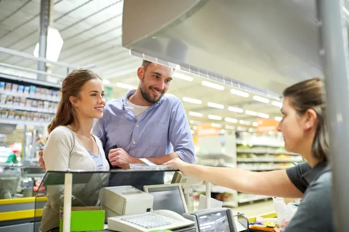 shopping, sale, consumerism, payment and people concept - happy couple buying food at grocery store or supermarket cash register and cashier taking money