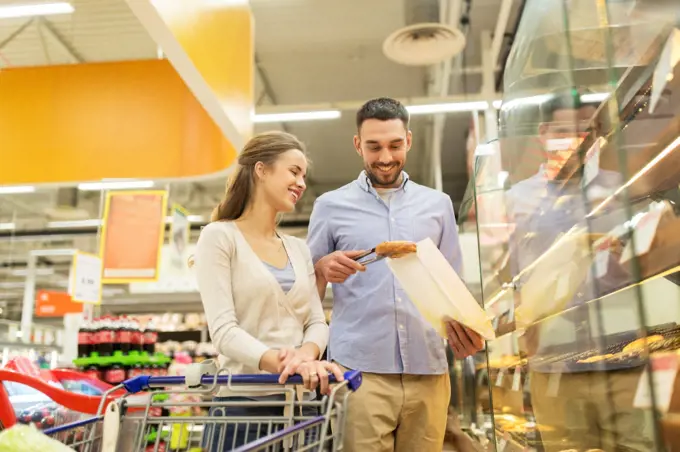 food, sale, consumerism and people concept - happy couple with shopping cart at grocery store or supermarket baking department buying buns or pies. happy couple with shopping cart at grocery store