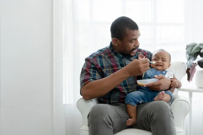 African toddler baby girl crying and dont want to eat food from her father who feeding with spoon while sitting on sofa at home. Little child care and relationship of dad and little daughter concept