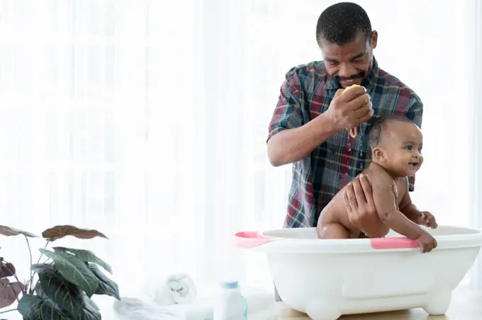 Happy African father with beard bathing adorable newborn baby daughter in bathtub at home. Dad cleaning his little kid girl with sponge in warm water. Child hygiene cleanliness care concept