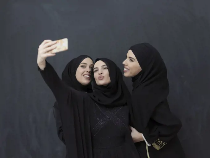 Portrait of Arab women wearing traditional clothes or abaya. Young muslim girls taking selfie in front of black chalkboard representing islamic arabic fashion and ramadan kareem concept