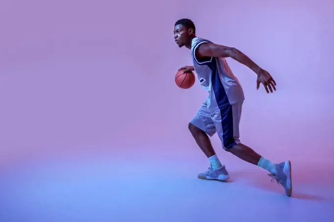 Basketball player practicing with ball in studio, neon background. Professional male baller in sportswear playing sport game, tall sportsman. Basketball player practicing with ball in studio