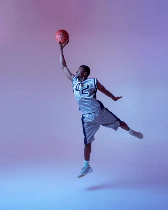 Basketball player with ball shows his skill in studio, jump in action, neon background. Professional male baller in sportswear playing sport game, tall sportsman. Basketball player with ball, jump in action