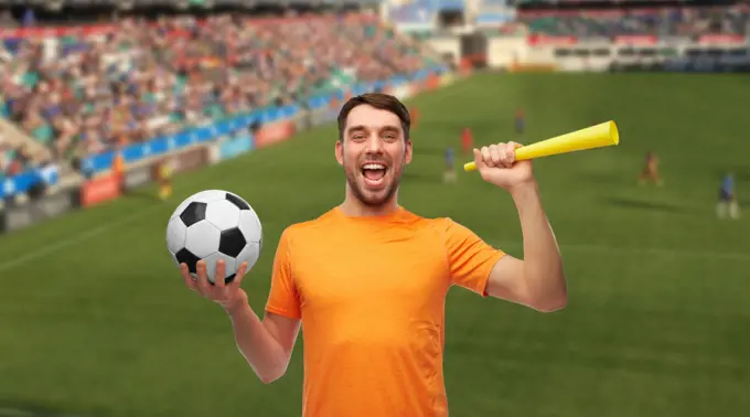 sport, leisure games and success concept - happy smiling man or football fan with soccer ball and vuvuzela over stadium background. male football fan with soccer ball and vuvuzela