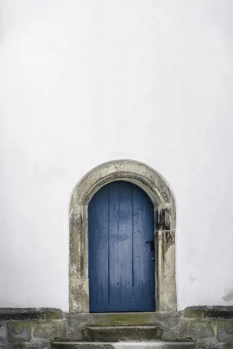 Medieval arched door. Aged wooden door in blue color with stone stairs and white wall. Just one door and an empty white house wall. Old blue entrance.