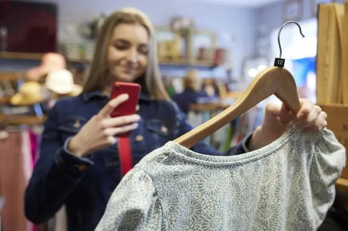 Young Woman Buying Used Sustainable Clothes From Second Hand Charity Shop Taking Picture Of Dress On Mobile Phone