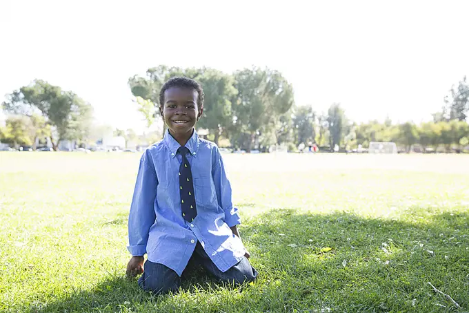 Portrait of boy kneeling on grass in sunlit park