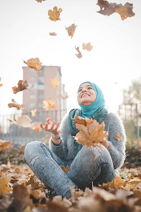 Young woman sitting in park throwing autumn leaves