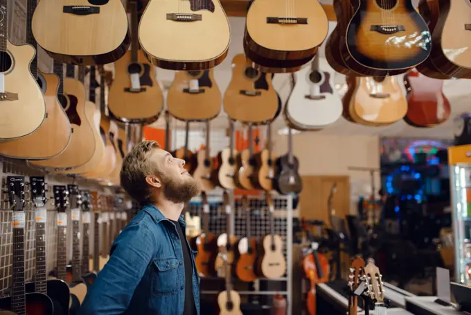 Bearded young guy choosing acoustic guitar in music store. Assortment in musical instruments shop, male musician buying equipment. Young guy choosing acoustic guitar in music store