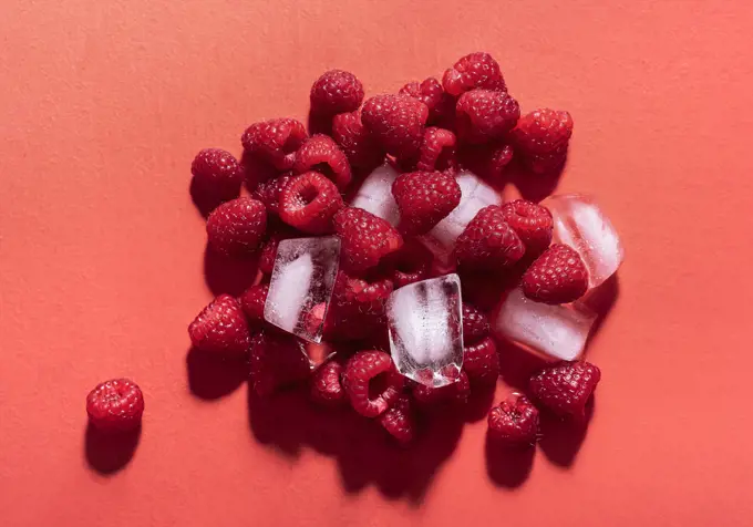 Fresh raspberries in a pile with ice cubes on a red background in sunlight. Above view of tasty summer fruits. Ingredients for berries lemonade.