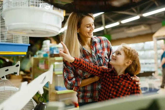 Mother with daughter choosing cage for bird at the showcase in pet store. Woman and little child buying equipment in petshop, accessories for domestic animals. Mother with daughter choosing cage in pet store