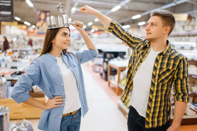 Young couple jokes with ladle and pan in houseware store. Man and woman buying home goods in market, family in kitchenware supply shop. Couple jokes with ladle and pan in houseware store