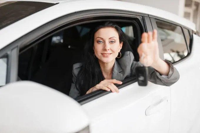 Happy woman shows the key to the new car in showroom. Female customer buying vehicle in dealership, automobile sale