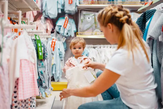 Mother with her little girl choosing clothes in kids store. Mom and child buying dress in supermarket together, family shopping