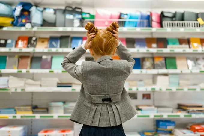 Little school girl at the shelf in stationery store, back view. Female child buying office supplies in shop, schoolchild in supermarket