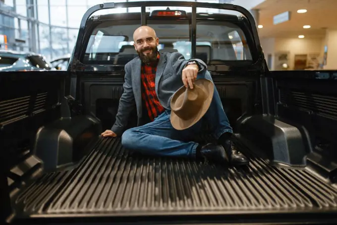 Smiling man poses in the back of new pickup truck in car dealership. Customer in vehicle showroom, male person buying transport, auto dealer business. Smiling man poses in the back of new pickup truck