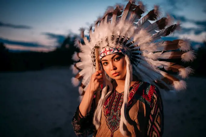 Portrait of young American Indian woman on sunset. Cherokee, Navajo, west native culture. Headdress made of feathers of wild birds. 