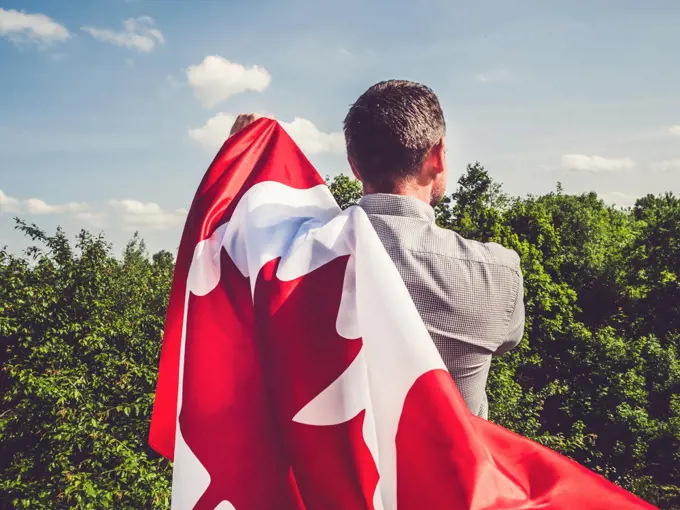 Attractive man holding Canadian Flag on blue sky background on a clear, sunny day. View from the back, close-up. National holiday concept. Attractive man holding Canadian Flag. National holiday