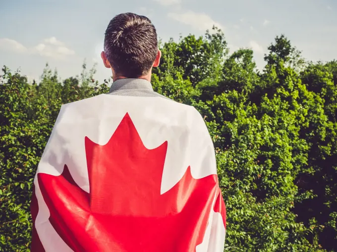 Attractive man holding Canadian Flag on blue sky background on a clear, sunny day. View from the back, close-up. National holiday concept. Attractive man holding Canadian Flag. National holiday