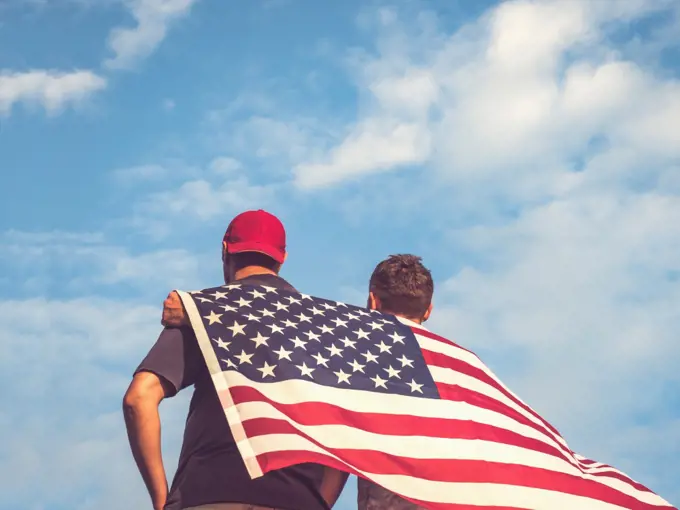 Two men holding Flag of the United States on blue sky background on a clear, sunny day. View from the back, close-up. National holiday concept. Two men holding Flag of the United States