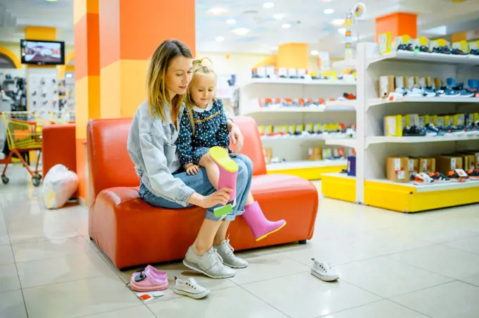 Mother and little daughter buying shoes in kid&rsquo;s store. Mom and adorable girl near the showcase in children&rsquo;s shop, happy childhood, family makes a purchase in market. Mother and little daughter buys shoes, kid&rsquo;s store