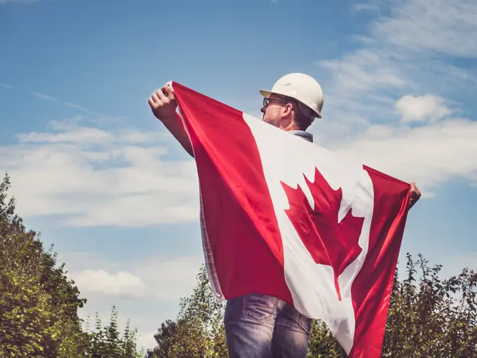 Handsome engineer, holding Canadian Flag in the park against the backdrop of green trees and the setting sun, looking into the distance. Close-up. Concept of labor and employment. Engineer, holding Canadian Flag in the park