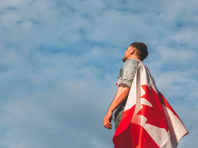 Attractive man in jeans and denim shirt holding a Canadian Flag against a clear, sunny, blue sky. View from the back, close-up. National holiday concept. Man holding a Canadian Flag. National holiday