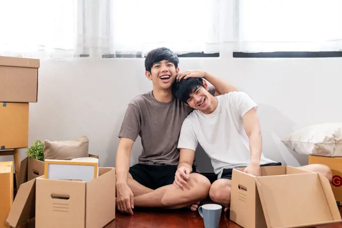 Portrait of young attractive happy asian couple man sitting at new home smiling to camera with carton package box storage to move in empty house. LGBTQ relationship and relocating concept