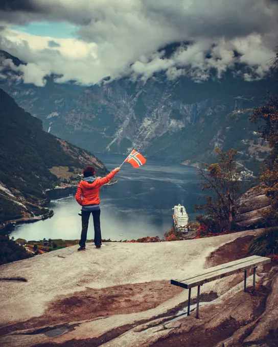 Female tourist enjoying scenic view over fjord Geirangerfjorden from Flydalsjuvet viewpoint, holding norwegian flag. Cruising vacation and travel.. Tourist over Geirangerfjord holds norwegian flag