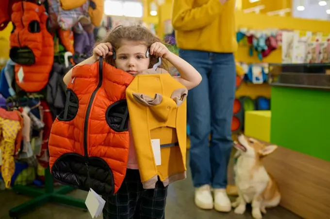 Little girl posing with trendy clothes for dog over pet shop interior. Happy mother and daughter shopping together buying new apparel for their corgi dog. Little girl posing with trendy clothes for dog over pet shop interior