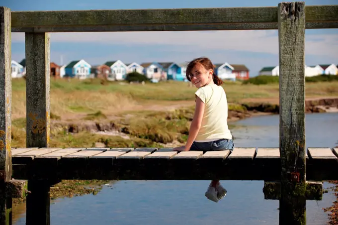 young girl sitting on foot bridge looking around at camera with beach huts in the background