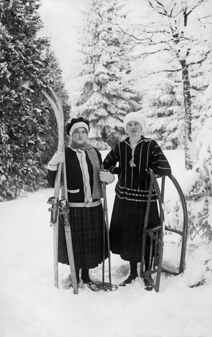 Two young women standing on snow holding sled and skis c.1910