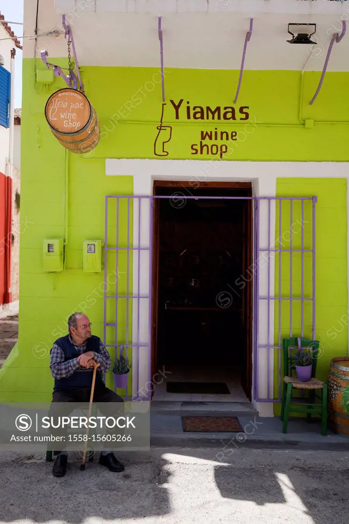 old man sits (supported by cane) in front of wine shop in greece