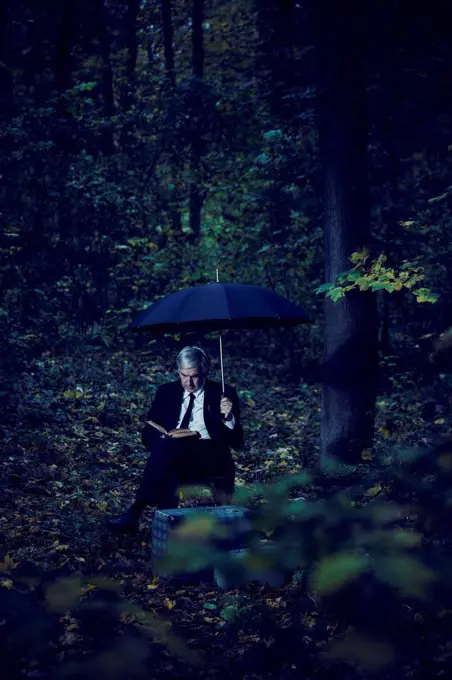 Man is sitting in the forest with umbrella book and two suitcases and is reading