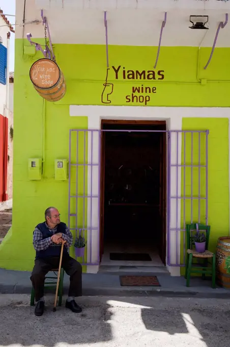 old man sits (supported by cane) in front of wine shop in greece